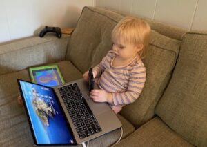 Toddler on a coach with a laptop, tablet, smartphone, and videogame controller